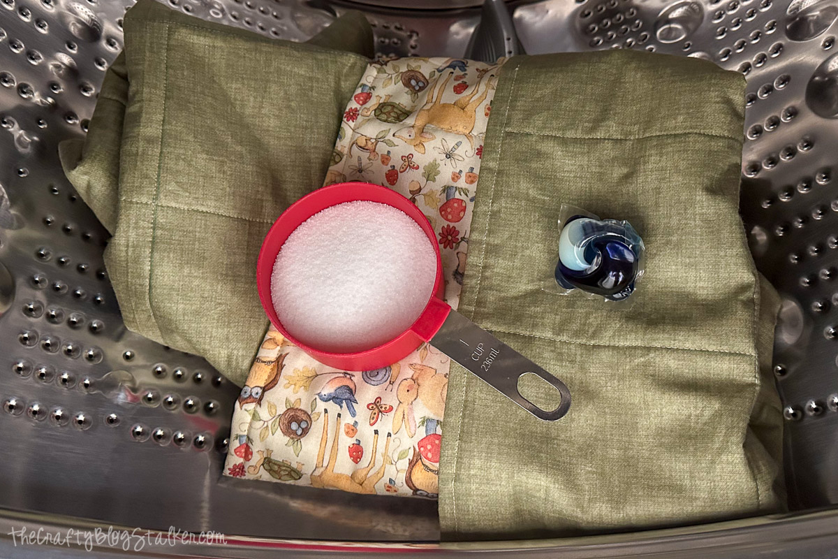 Baby blanket placed in a washing machine drum with a measuring cup of salt and a detergent pod on top before first wash.