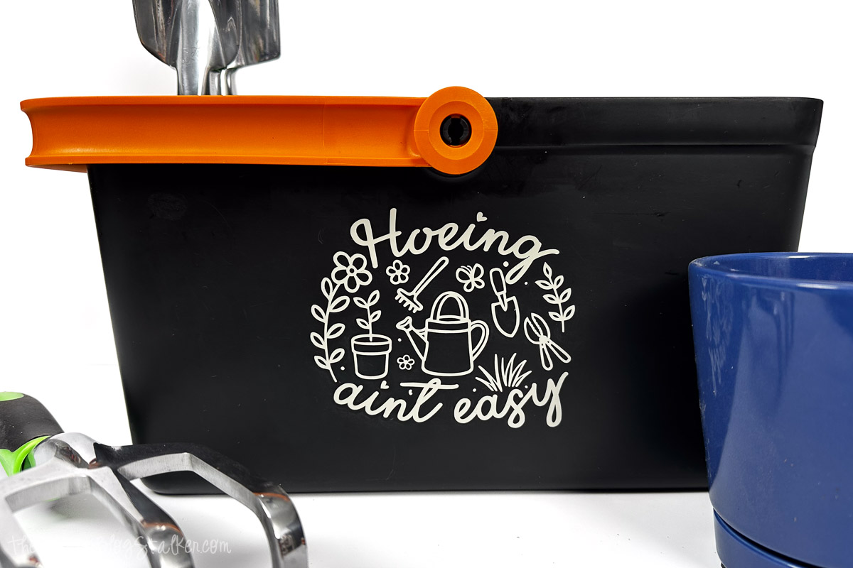 Black garden bucket with orange handle featuring a gardening design and phrase, surrounded by hand tools and stacked plant pots.