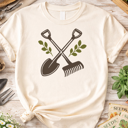 Flat lay of a T-shirt featuring crossed shovel and rake graphic with leafy accents, styled on a rustic table with gardening tools and seed packets.