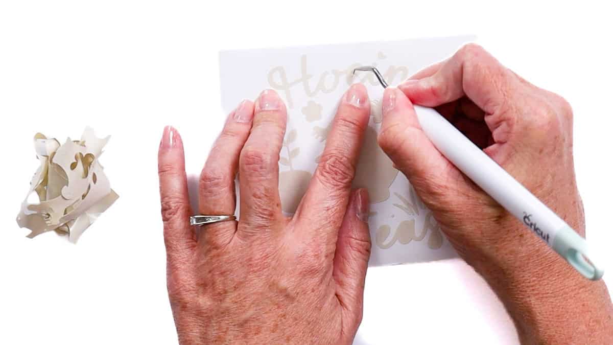 Hands use a weeding tool to remove excess vinyl from a small cut design while scraps sit to the side.