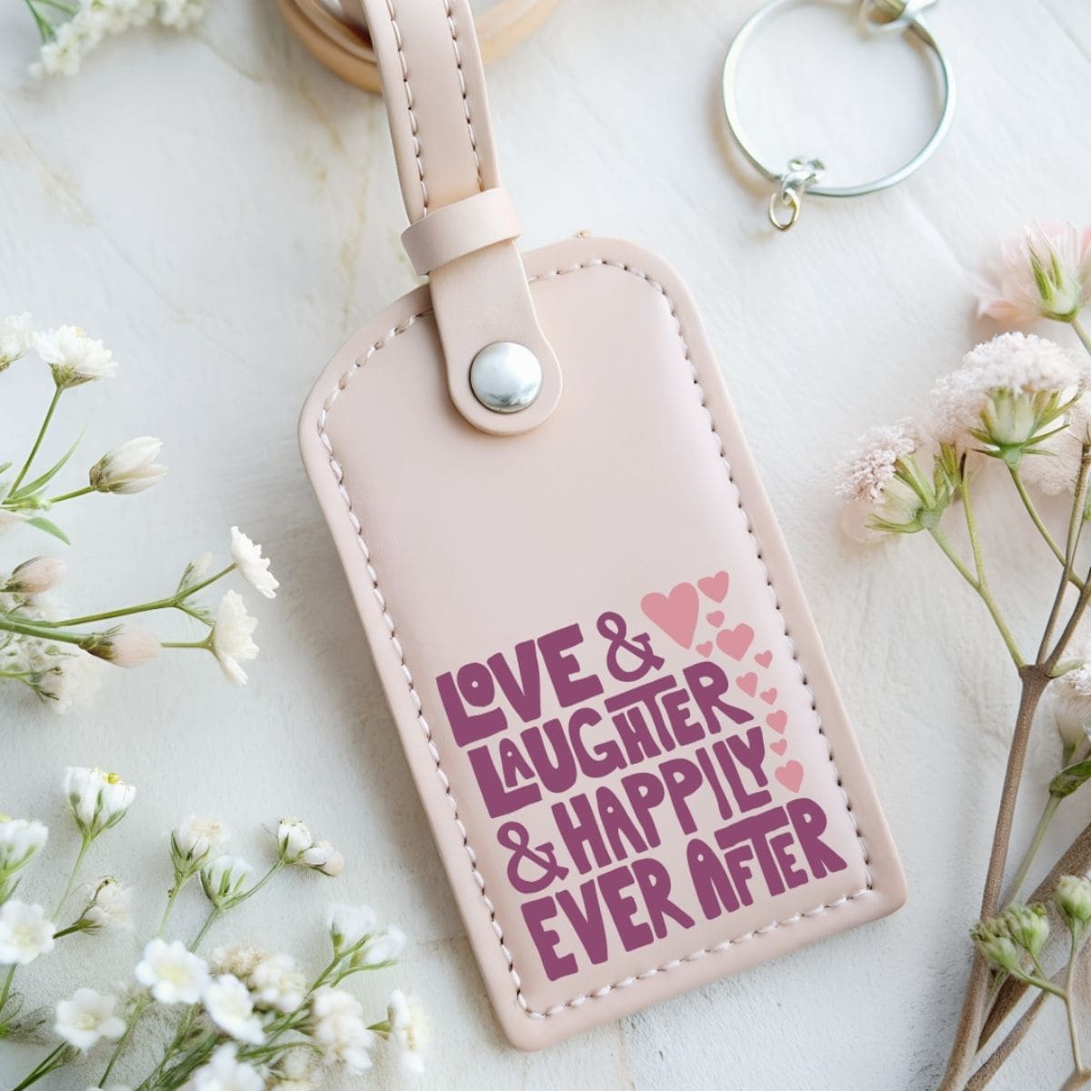 Blush pink luggage tag with the phrase Love & Laughter & Happily Ever After in purple vinyl, styled with small hearts and surrounded by delicate flowers.