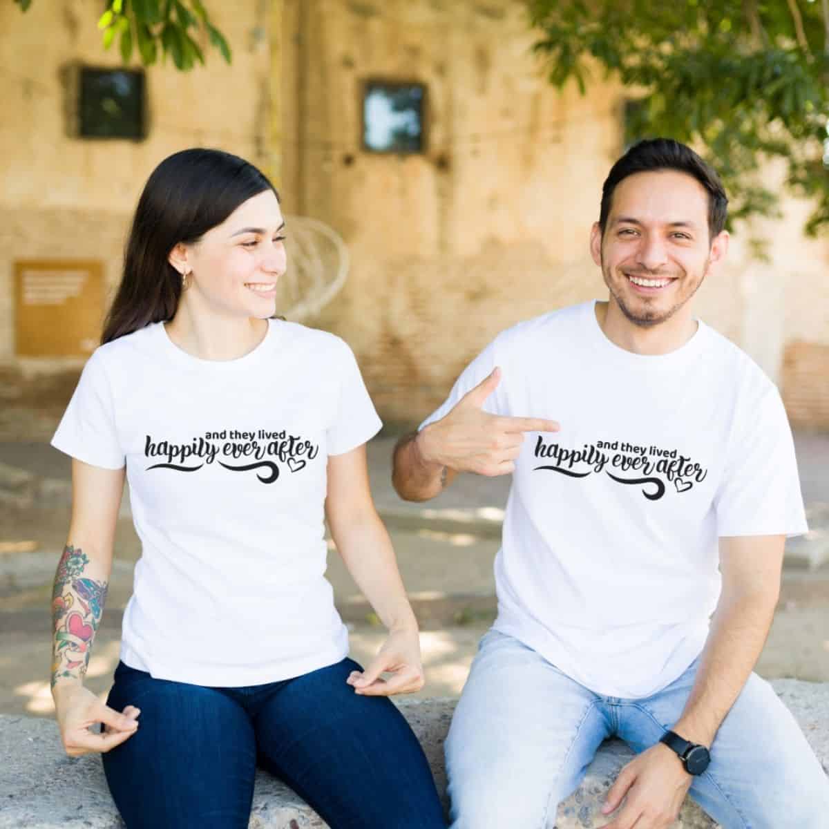 Couple wearing matching white T-shirts with the phrase and they lived happily ever after, sitting outdoors and smiling at each other.
