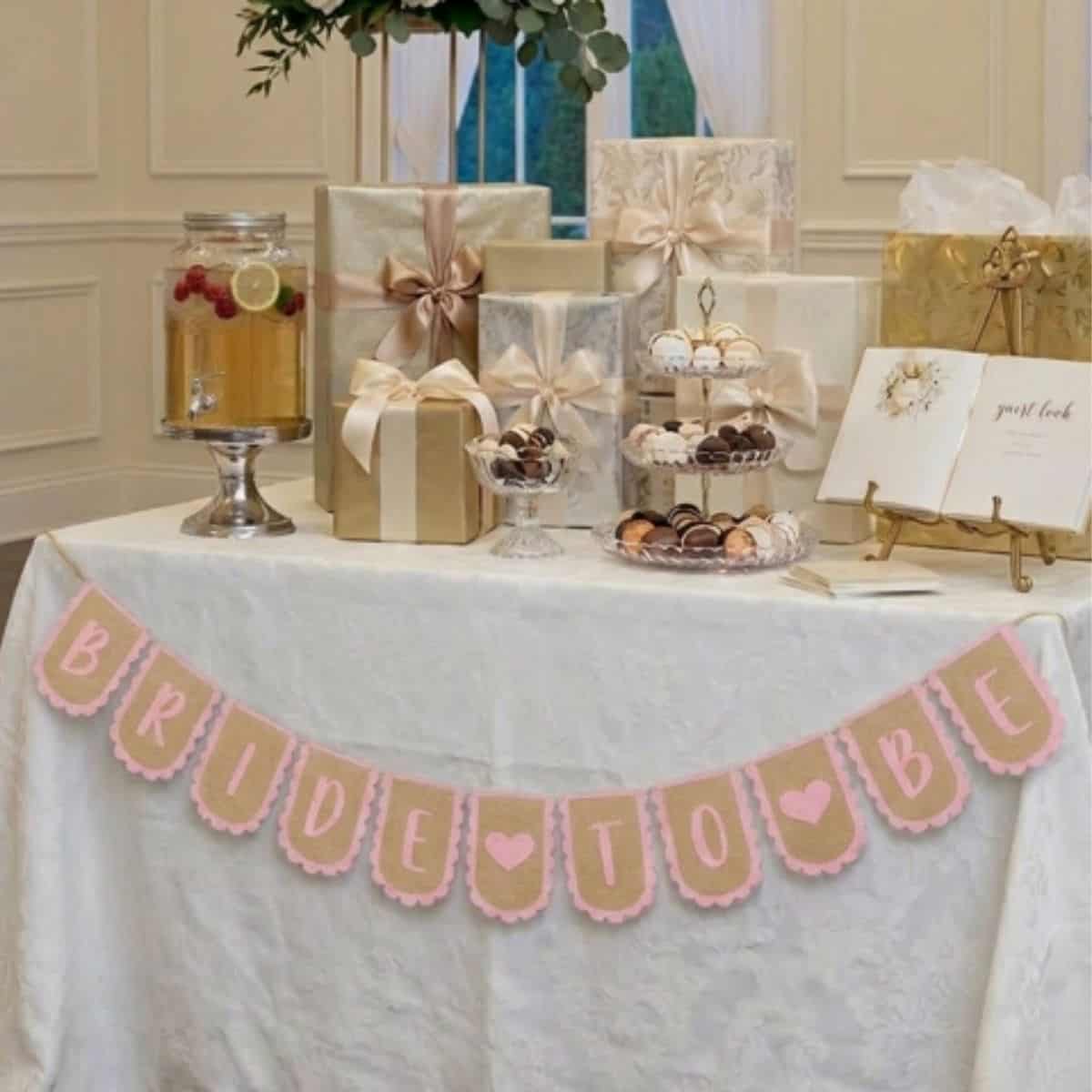 Bridal shower gift table with wrapped presents, desserts, and a pink and gold Bride to Be banner hanging across the front.