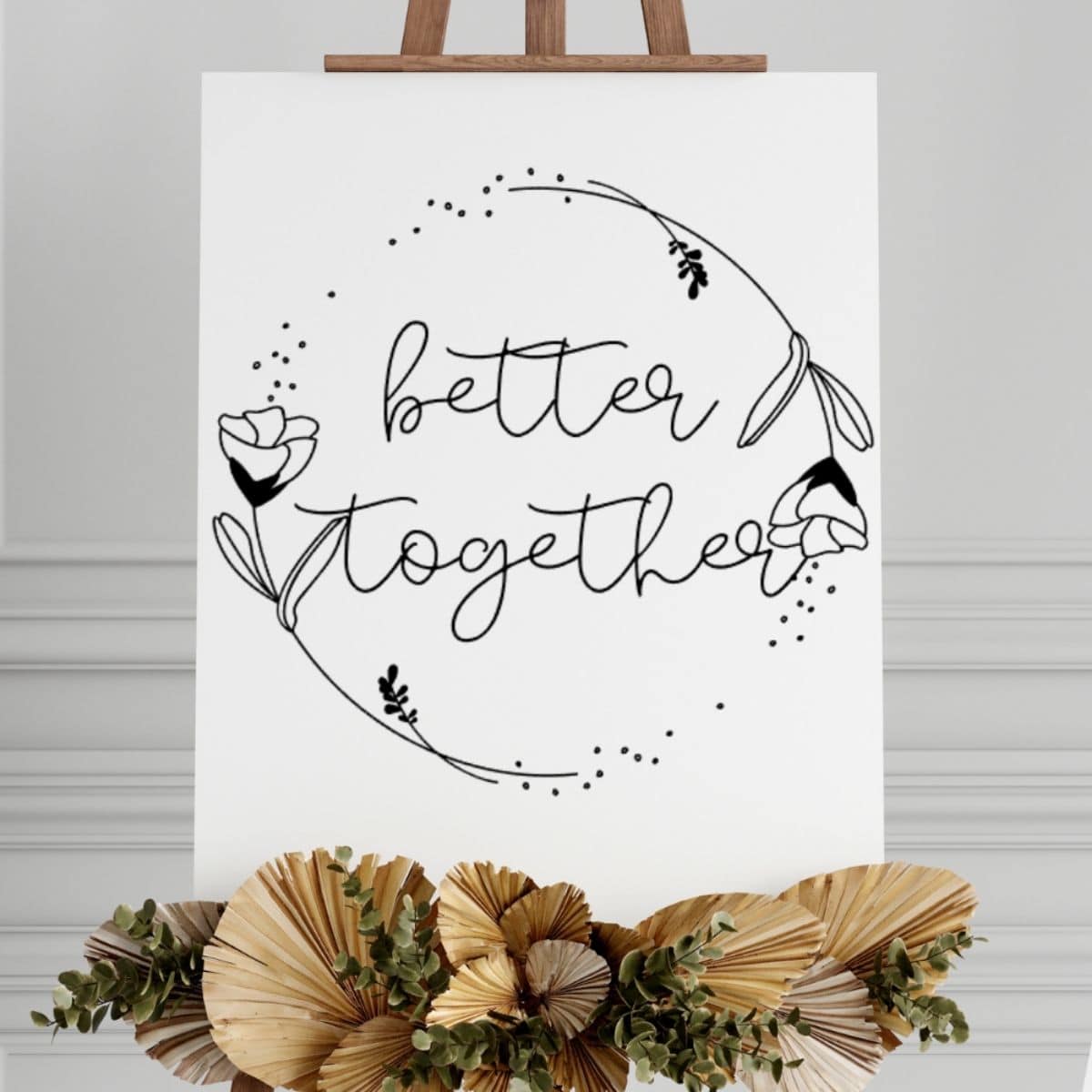 Wedding sign on an easel with the words better together inside a floral wreath design, decorated with dried palm leaves and greenery.