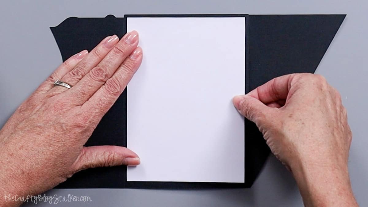 Hands placing a white cardstock panel inside a folded black card base while assembling a layered bride and groom wedding card.