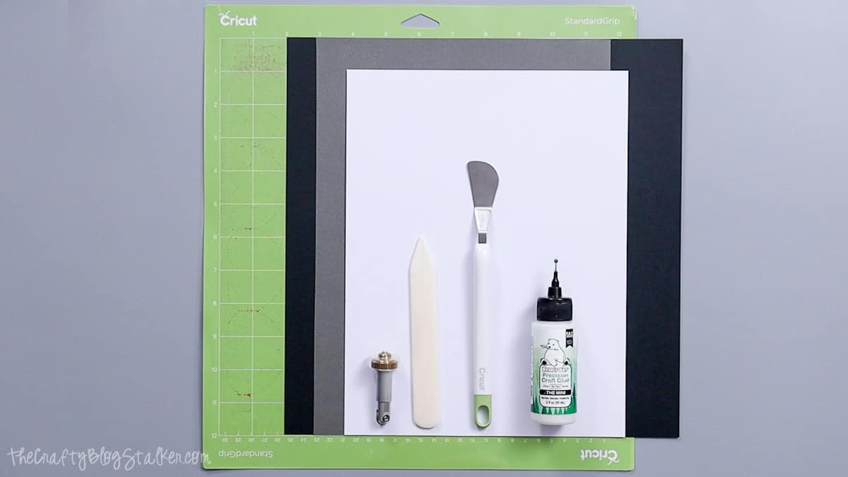 Cricut mat with cardstock, brayer, bone folder, spatula tool, and craft glue arranged as supplies for making a layered paper card.