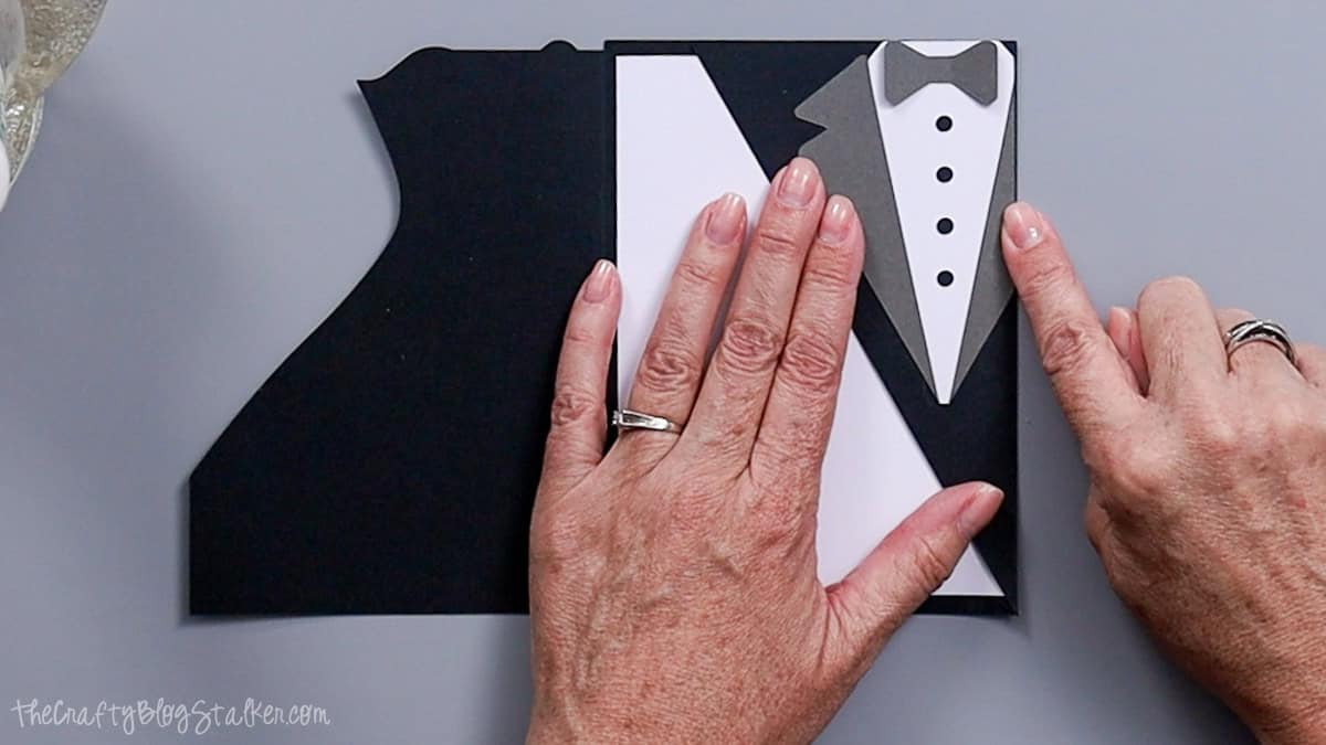 Hands pressing a layered cardstock tuxedo with bow tie and buttons onto a black card base while assembling a bride and groom wedding card.
