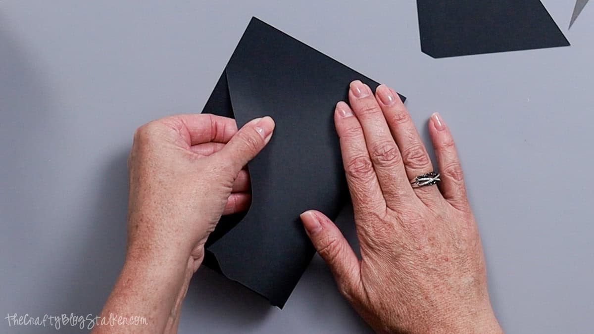Hands folding a black cardstock card base along the scored line to begin assembling a layered bride and groom wedding card.