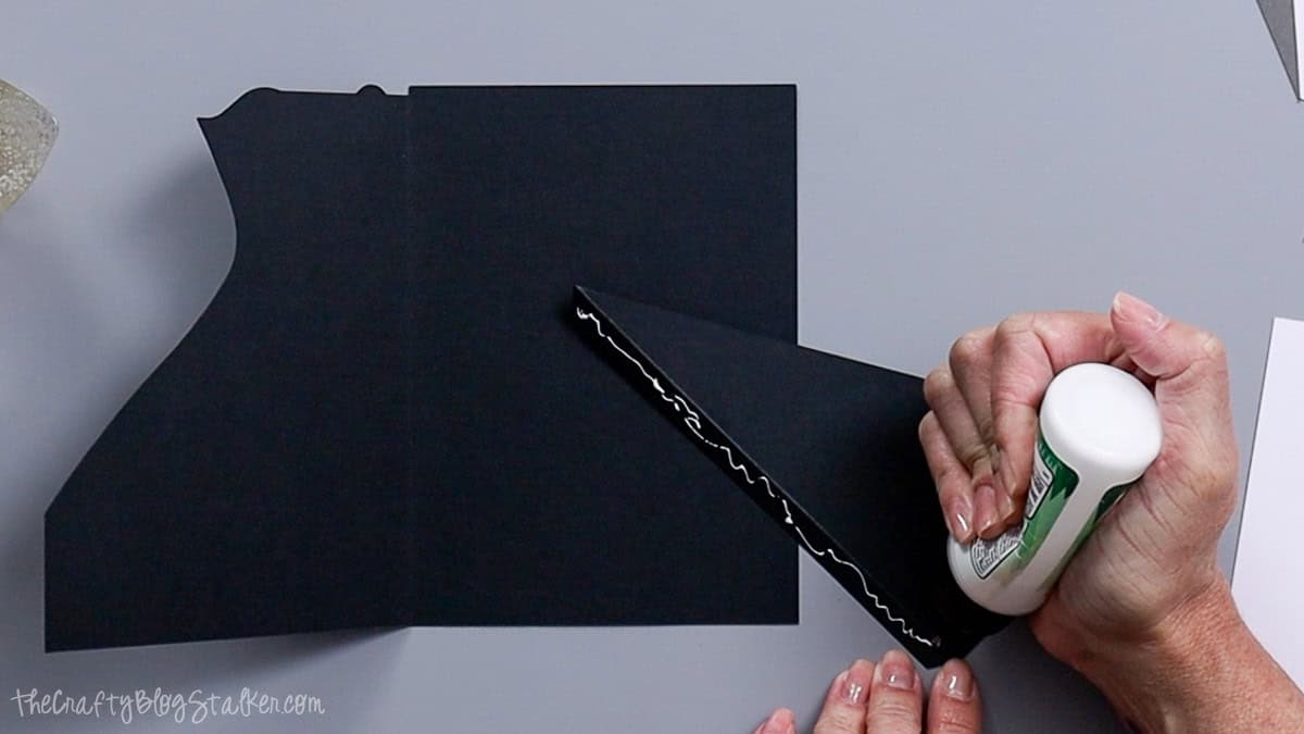 Hands applying craft glue to a triangular cardstock flap while assembling a black bride and groom themed paper card.