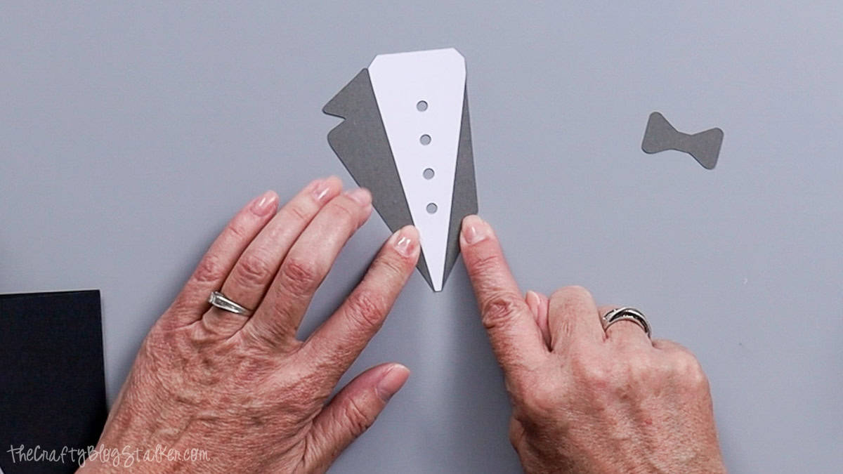 Hands aligning layered cardstock pieces to assemble a groom's tuxedo with buttons and bow tie for a handmade wedding card.