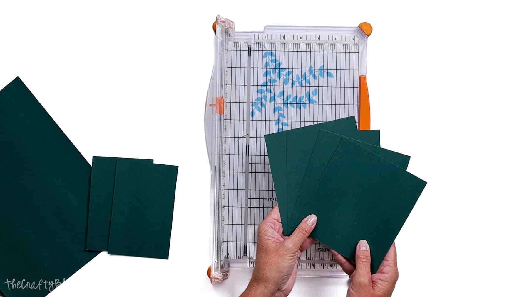 Hands holding several cut cardstock pieces next to a paper trimmer with grid lines and sliding blade, showing multiple evenly trimmed rectangles.