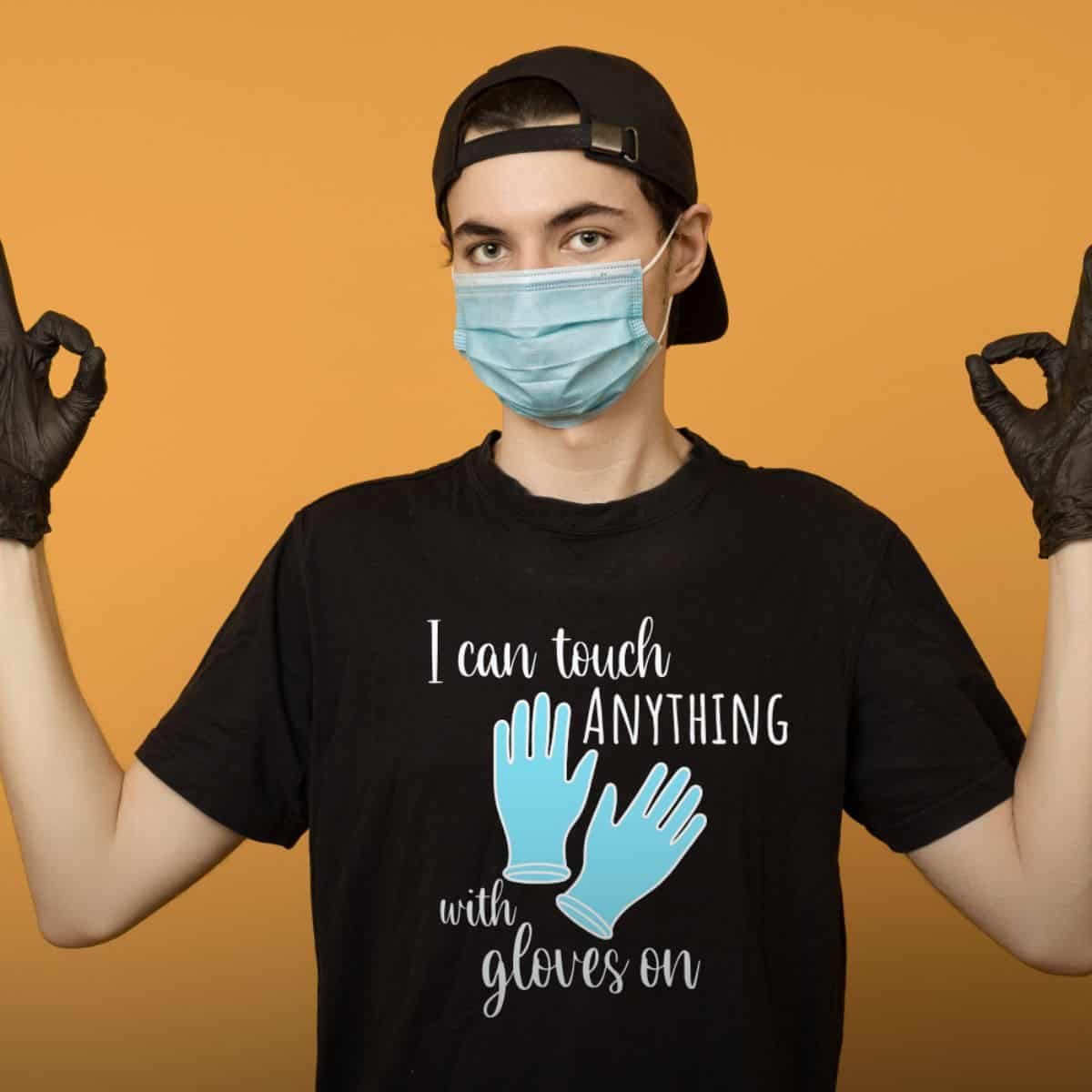 Person wearing a face mask and black gloves poses with hands raised, wearing a black T-shirt that says 