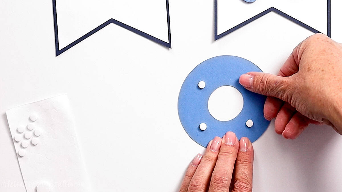Hands applying adhesive foam dots to the back of a blue letter O piece for a layered birthday banner.