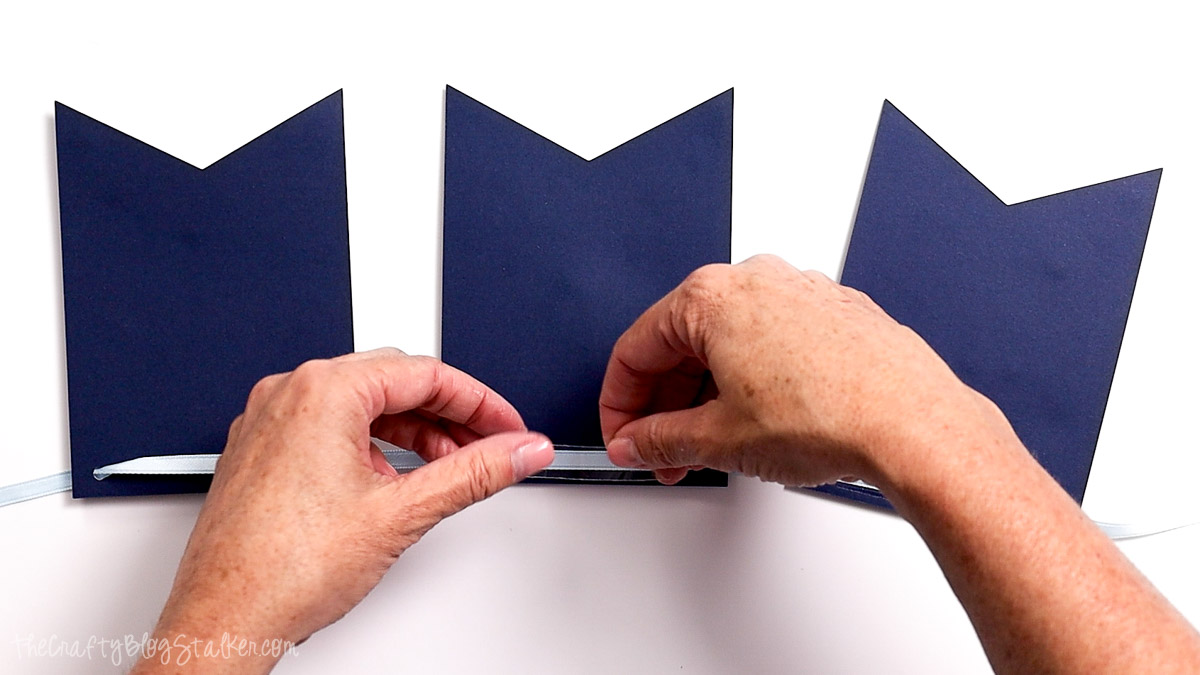 Hands applying double-sided tape to the back of blue banner pennants to attach ribbon for a birthday banner.