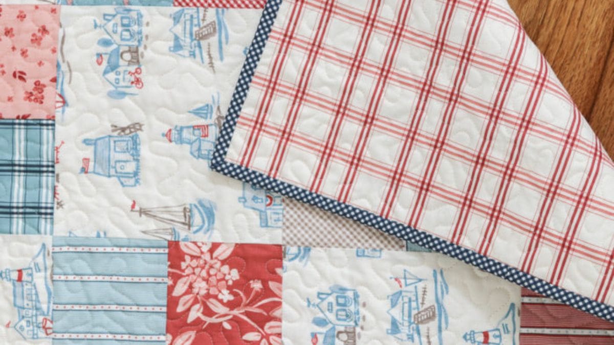 Patchwork quilt with red, blue, and white fabric squares featuring coastal prints and stripes, with a folded corner showing plaid backing and quilted stitching