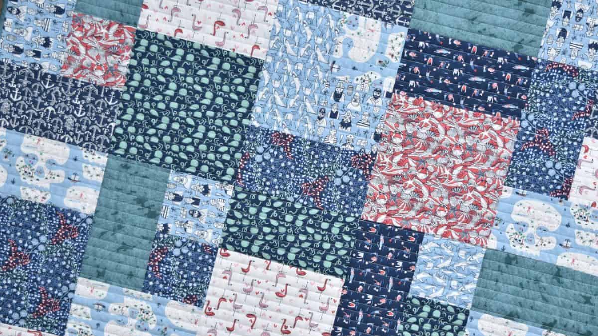 Patchwork quilt made from blue, teal, and red patterned fabric squares arranged in a grid with visible stitching lines and mixed prints including animals and florals