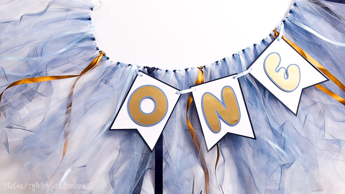 Blue tulle and ribbon garland with white pennant flags spelling ONE in gold letters, styled as a first birthday high chair banner.