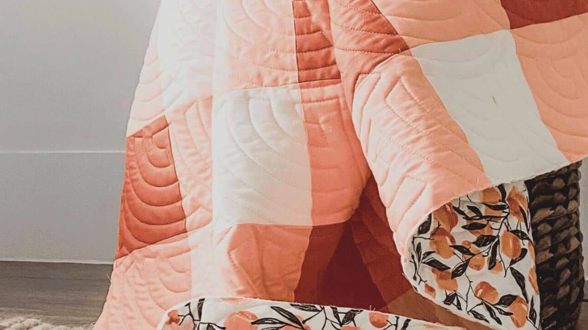 Close-up of a peach and white gingham quilt with curved quilting stitches draped over a basket, showing a floral backing fabric at the edge