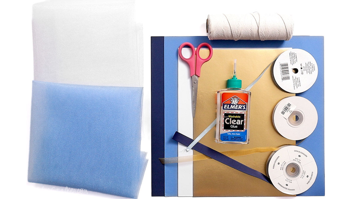 Craft supplies for a first birthday banner including tulle, cardstock, scissors, glue, ribbon, twine, and foam sheets arranged on a table.