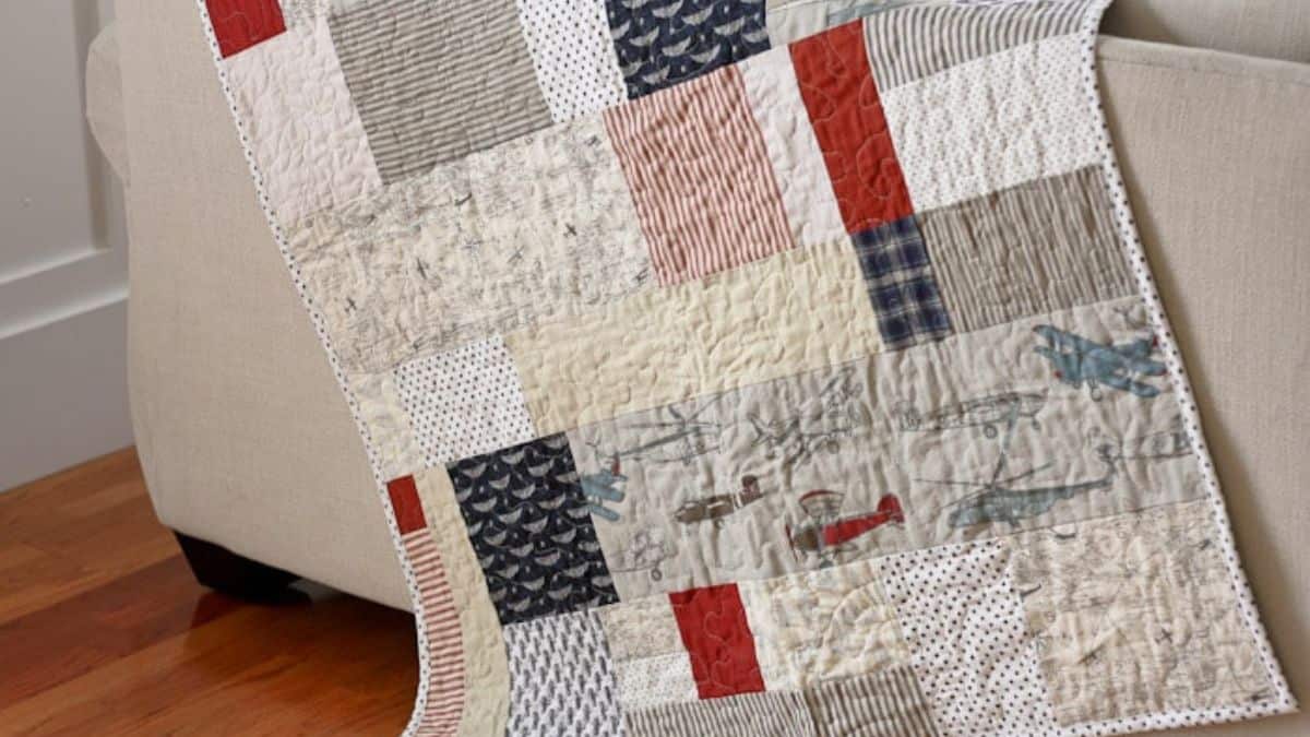 Patchwork quilt with neutral and red fabric squares draped over a chair, featuring airplane prints and textured quilting on a hardwood floor