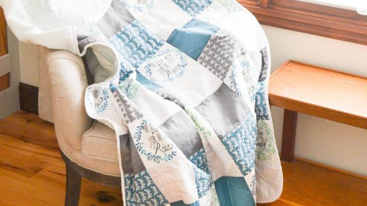 Blue, gray, and white patchwork quilt draped over an armchair near a wooden bench, showing mixed patterned squares and soft quilted texture