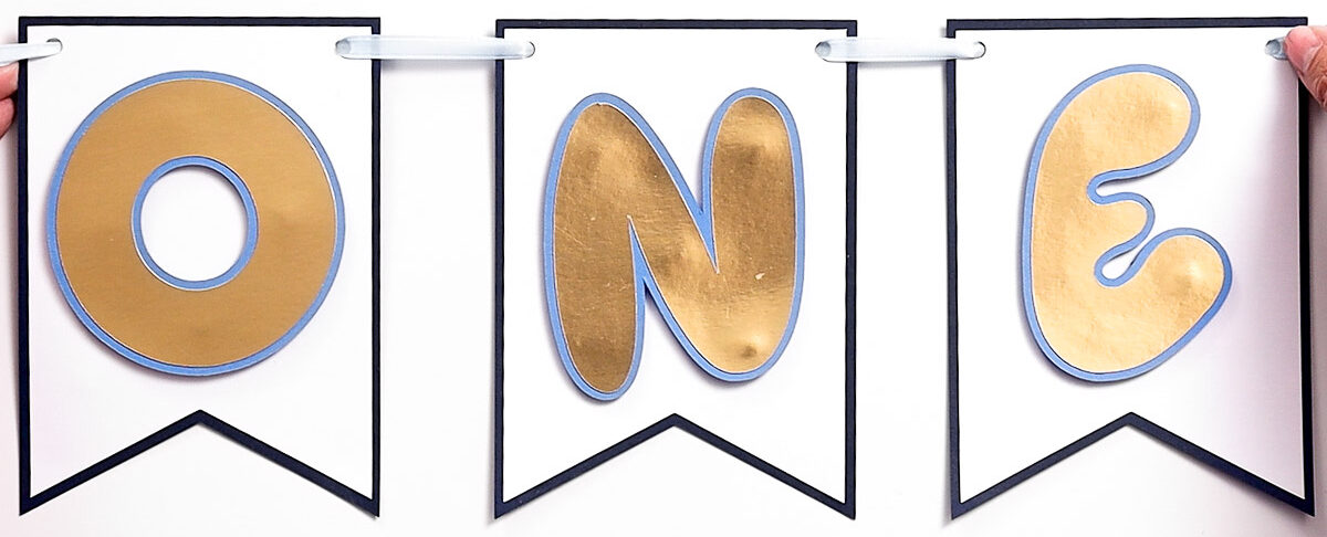 Finished ONE banner with gold foil letters on white pennants outlined in blue, strung together with ribbon.