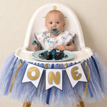 Baby in a high chair with a “ONE” banner, eating a smash cake with blue frosting during a first birthday celebration.