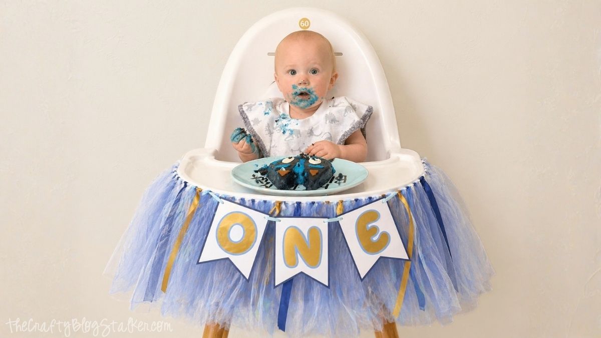 Baby in a high chair with blue cake on face, wearing a bib, with a blue tulle high chair banner and ONE pennant flags for a first birthday.