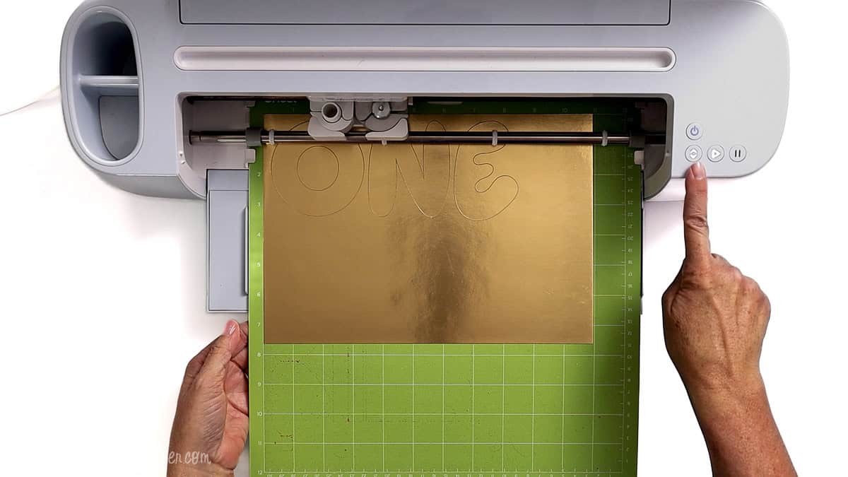 Cricut machine cutting letters on gold foil cardstock placed on a green cutting mat while a hand presses the start button.