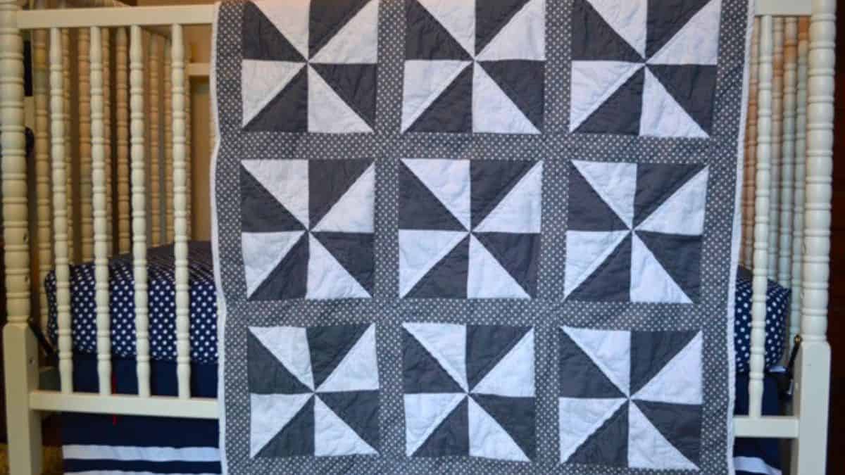 Gray and white pinwheel quilt with repeating geometric blocks hanging over a crib, showing a classic pinwheel pattern and stitched quilting lines