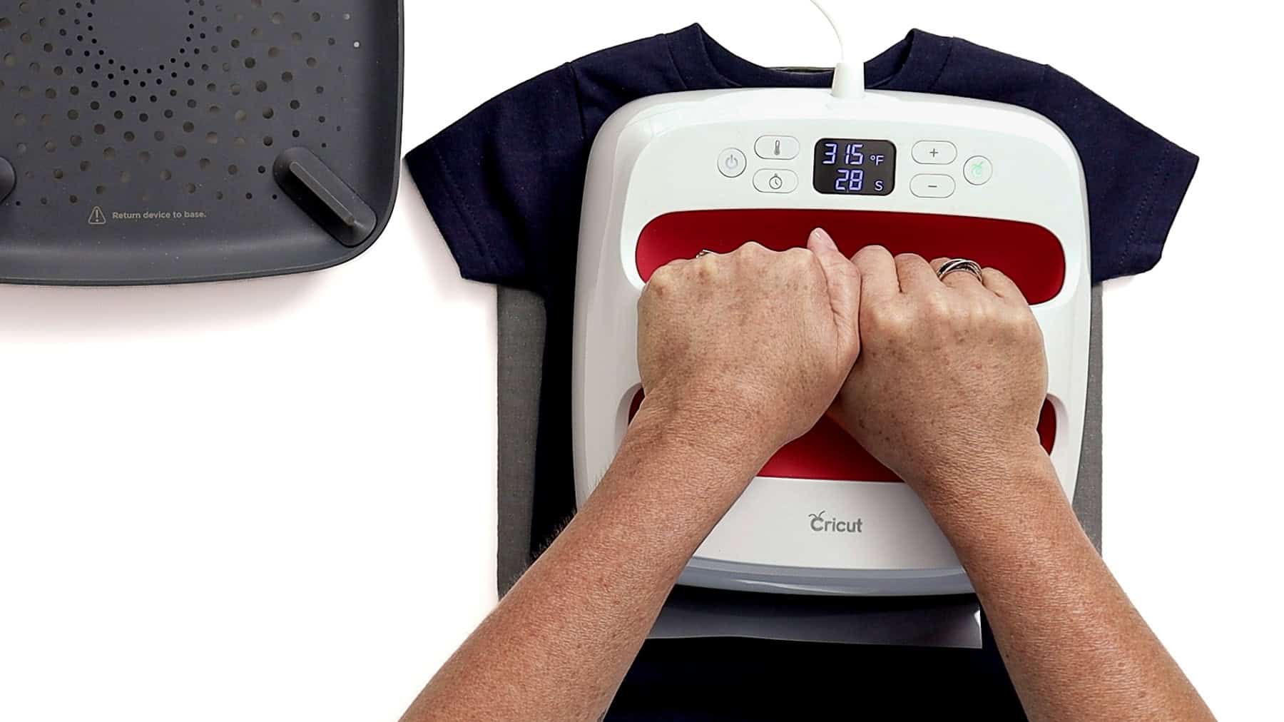 Hands pressing a Cricut EasyPress onto a navy toddler shirt to apply iron-on vinyl, with heat set to 315°F for 20 seconds
