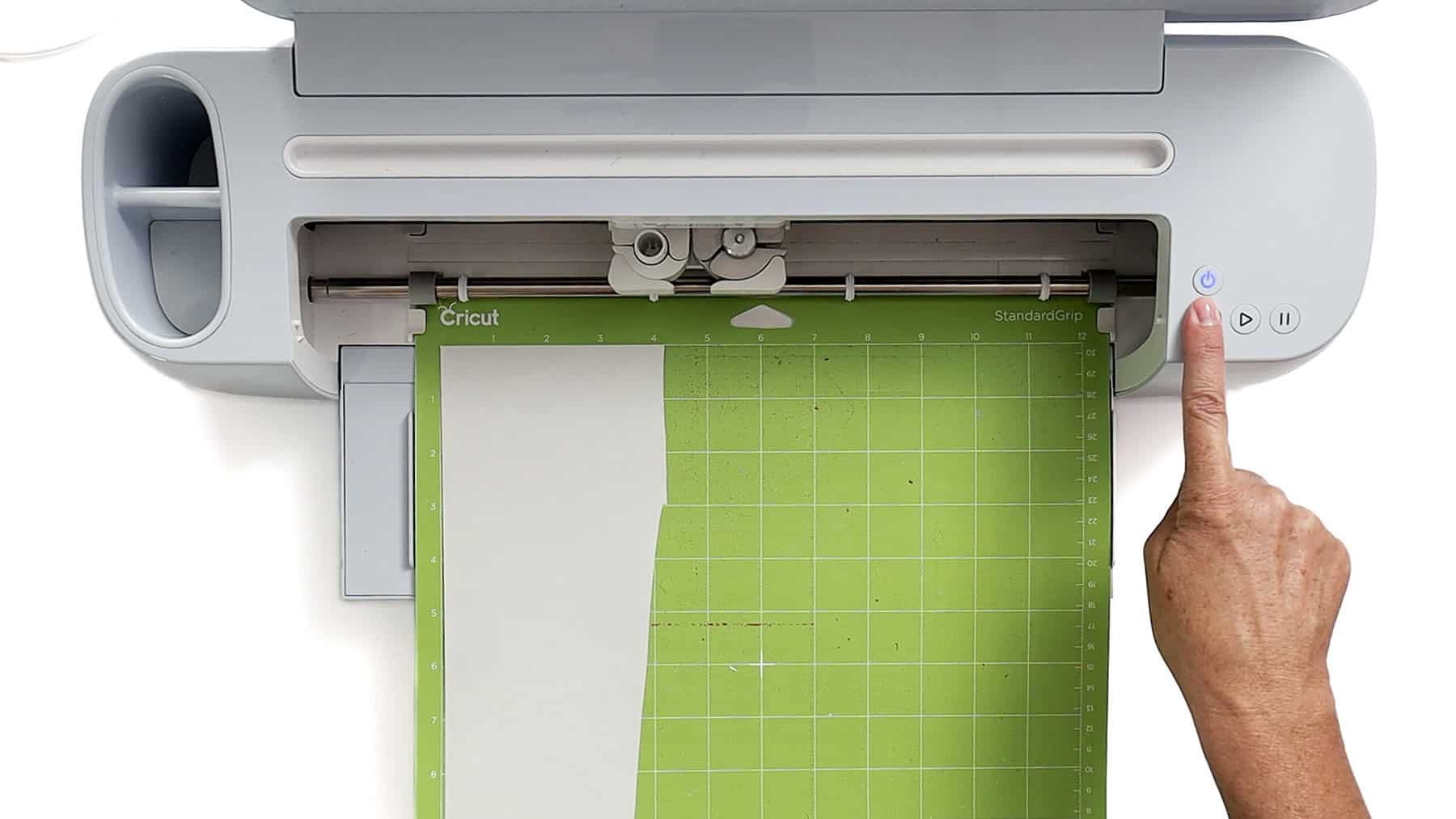 Cricut cutting machine with green StandardGrip mat loaded and a hand pressing the power button to start cutting
