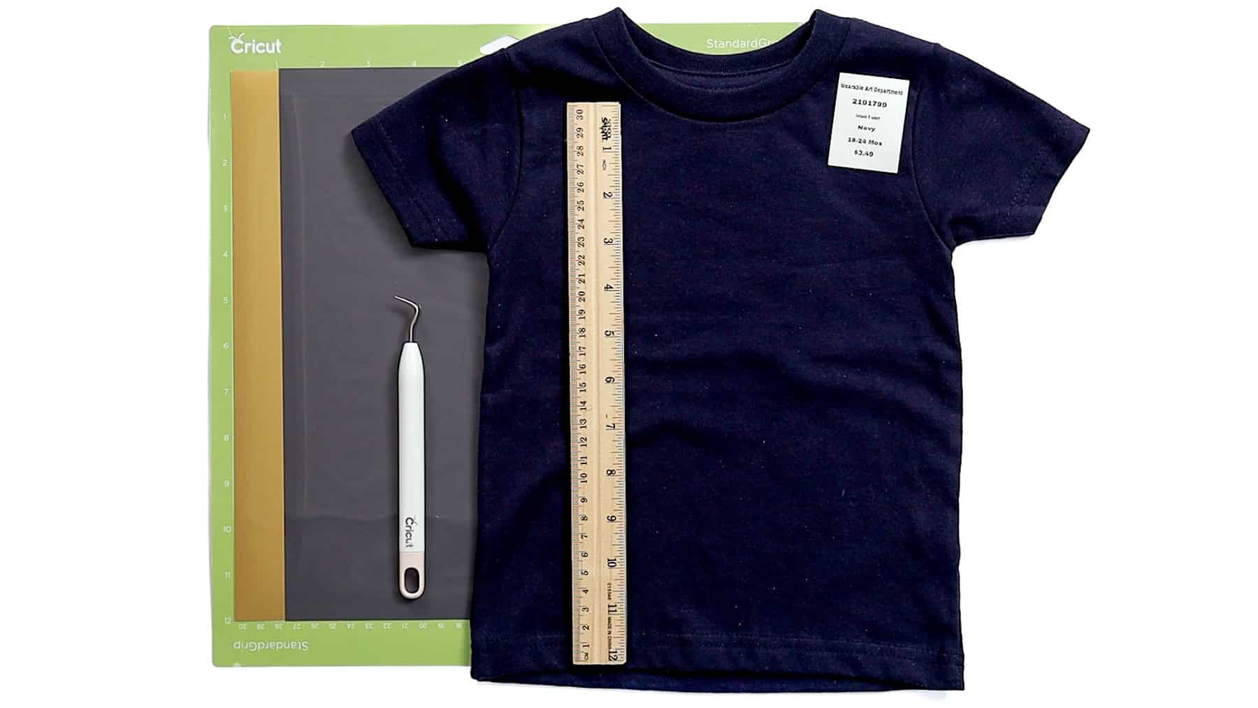 Navy toddler T-shirt with a ruler placed down the center, alongside a Cricut cutting mat and weeding tool for measuring and crafting setup