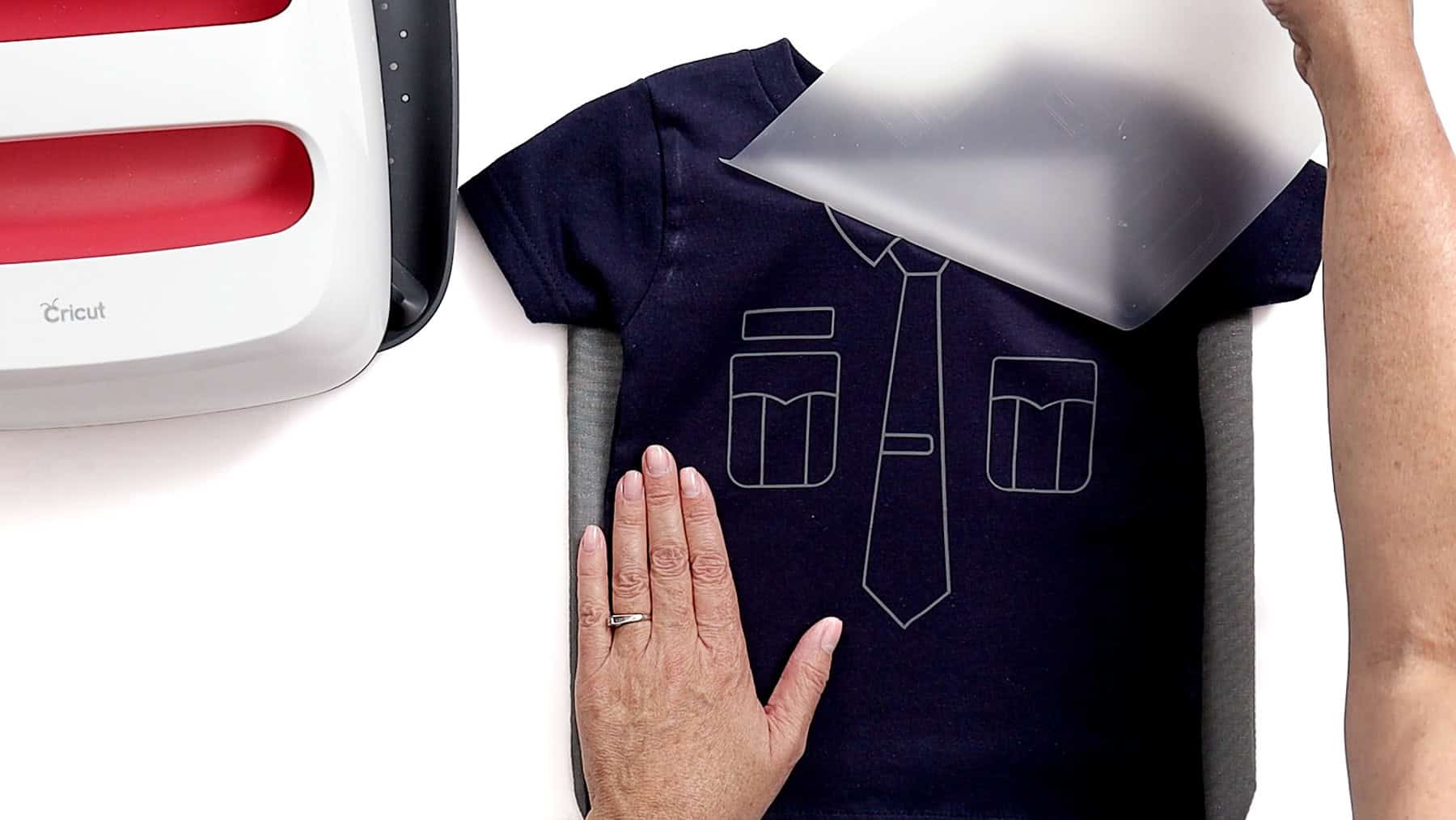 Hand peeling clear carrier sheet from a navy toddler shirt, revealing a white police uniform iron-on vinyl design after heat pressing
