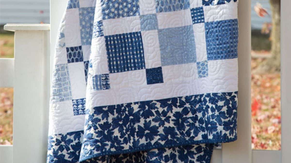 Blue and white patchwork quilt with mixed patterned squares and floral border draped over a porch railing, showing detailed quilting stitches