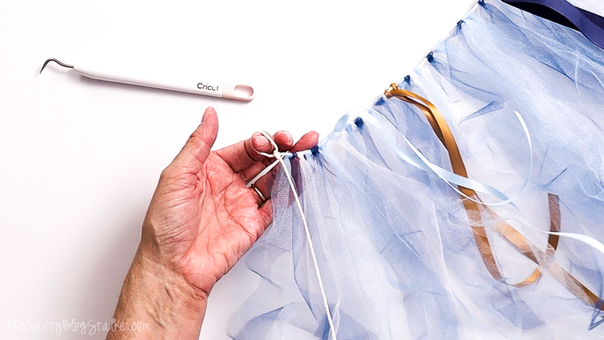 Hand tying a loop in string at the end of a blue tulle and ribbon garland, with a Cricut weeding tool nearby.