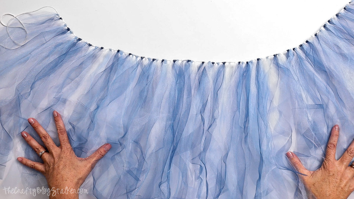 Hands spreading out a full tulle garland made of light blue and white strips tied along a string for a birthday banner decoration.