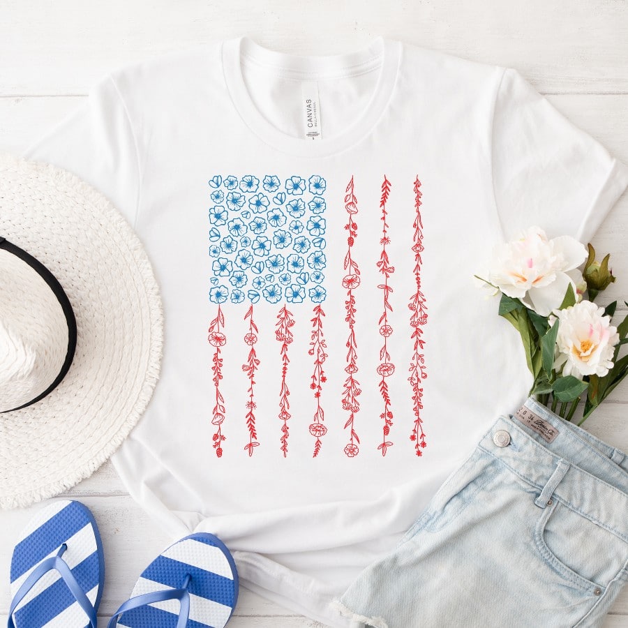 White T-shirt with a floral American flag design styled with denim shorts, blue flip flops, flowers, and a straw hat.