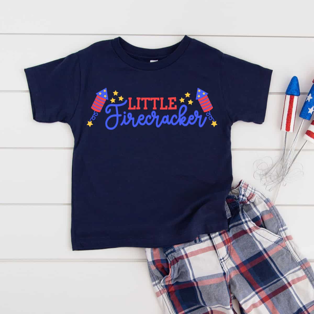 Navy Little Firecracker kids T-shirt with patriotic graphics styled with plaid shorts and red, white, and blue fireworks props.