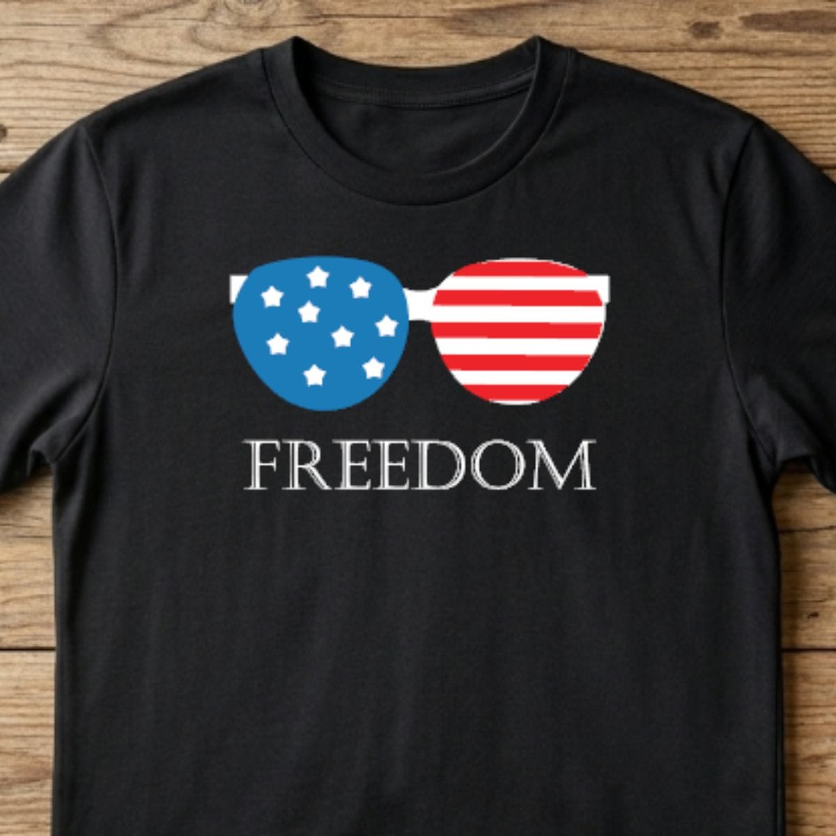 Black T-shirt with patriotic sunglasses graphic featuring stars and stripes and the word Freedom on a rustic wood background.