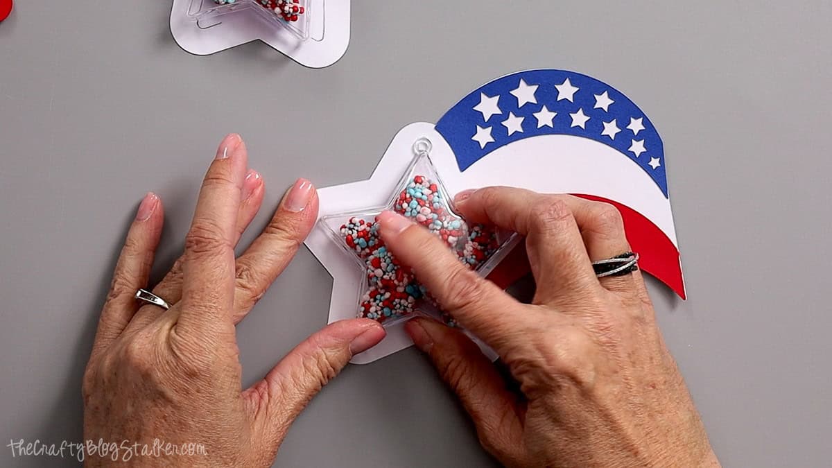 Hands attaching a clear star shaker ornament filled with candy to a patriotic red, white, and blue paper party favor.