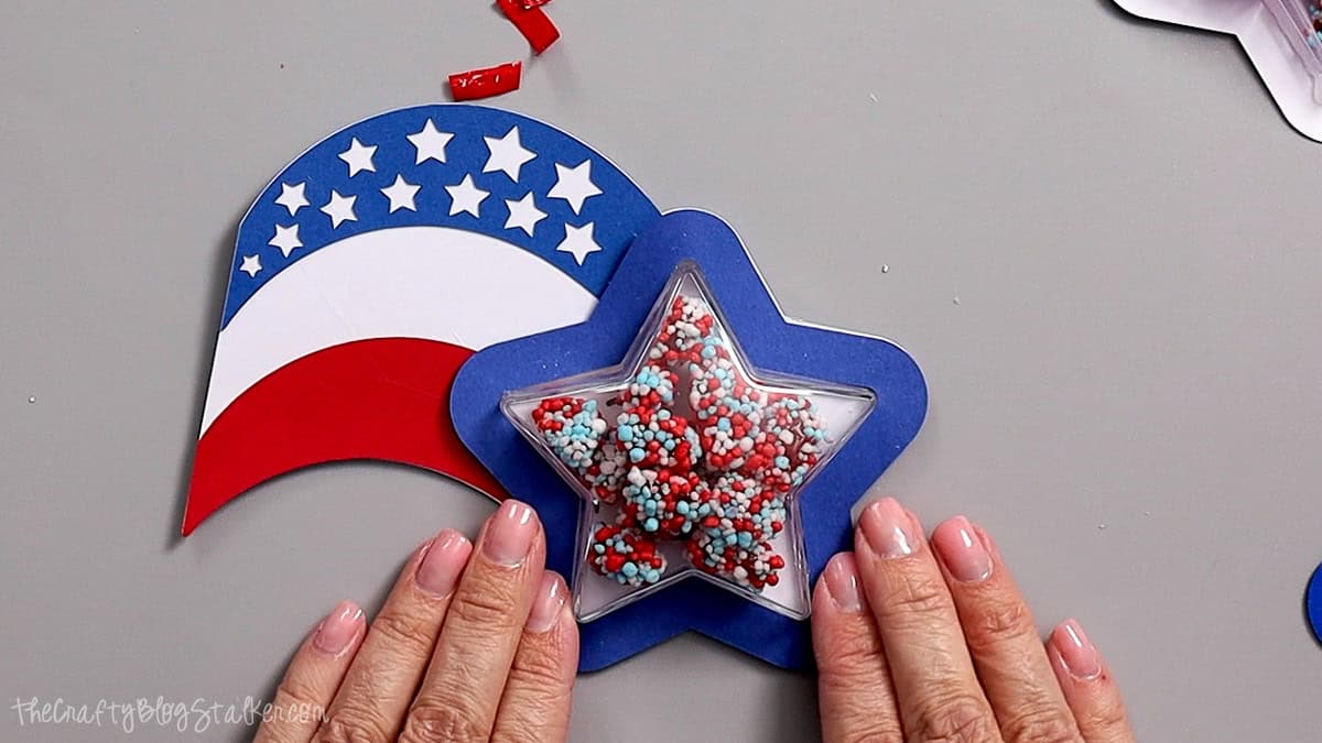 Hands holding a patriotic shaker party favor with a clear star filled with red, white, and blue candy sprinkles on layered cardstock.
