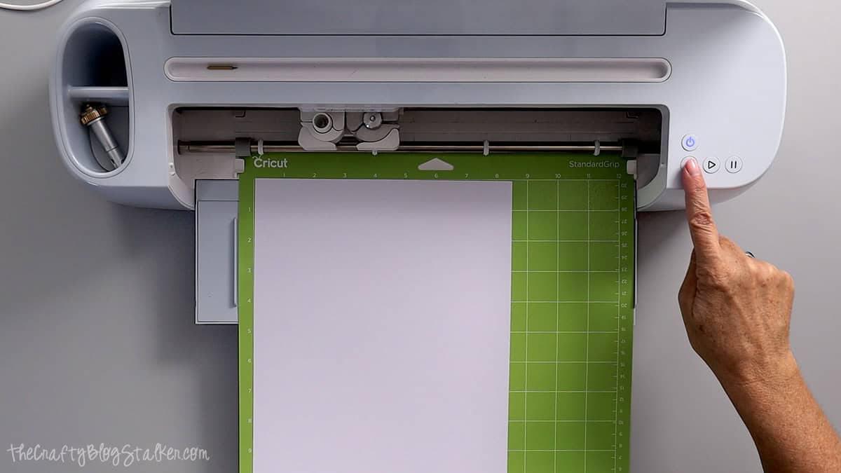 Hand pressing the Cricut machine start button with white cardstock loaded on a green StandardGrip cutting mat.