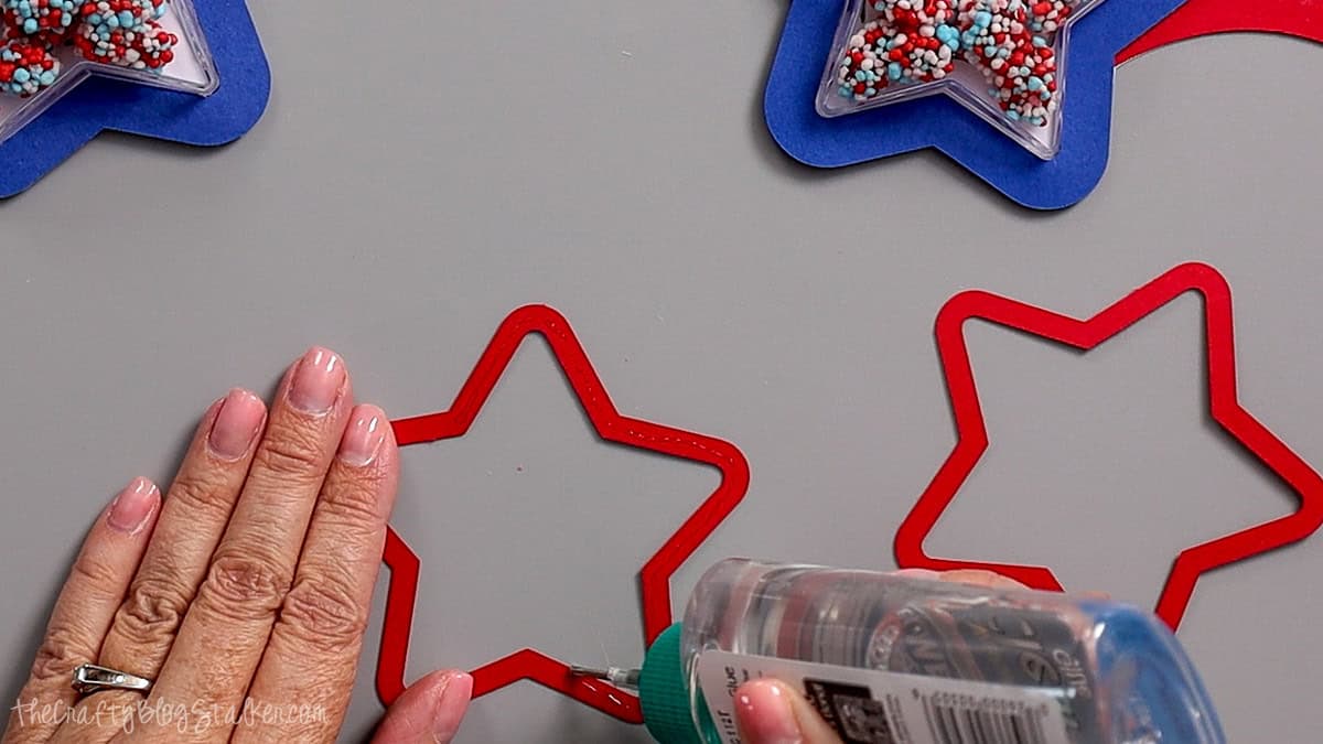 Hands applying glue to a red star cardstock frame while assembling patriotic shaker party favors with candy-filled star containers.