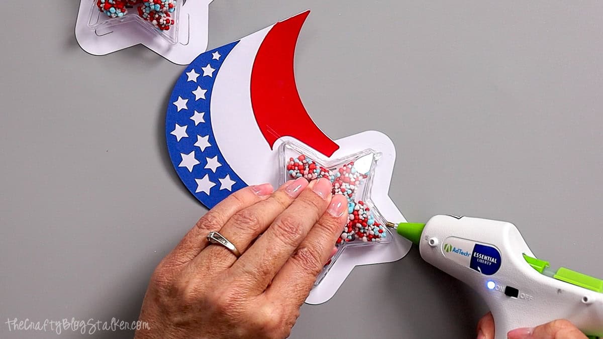 Hands using a hot glue gun to attach a candy-filled star ornament to a patriotic paper party favor craft.