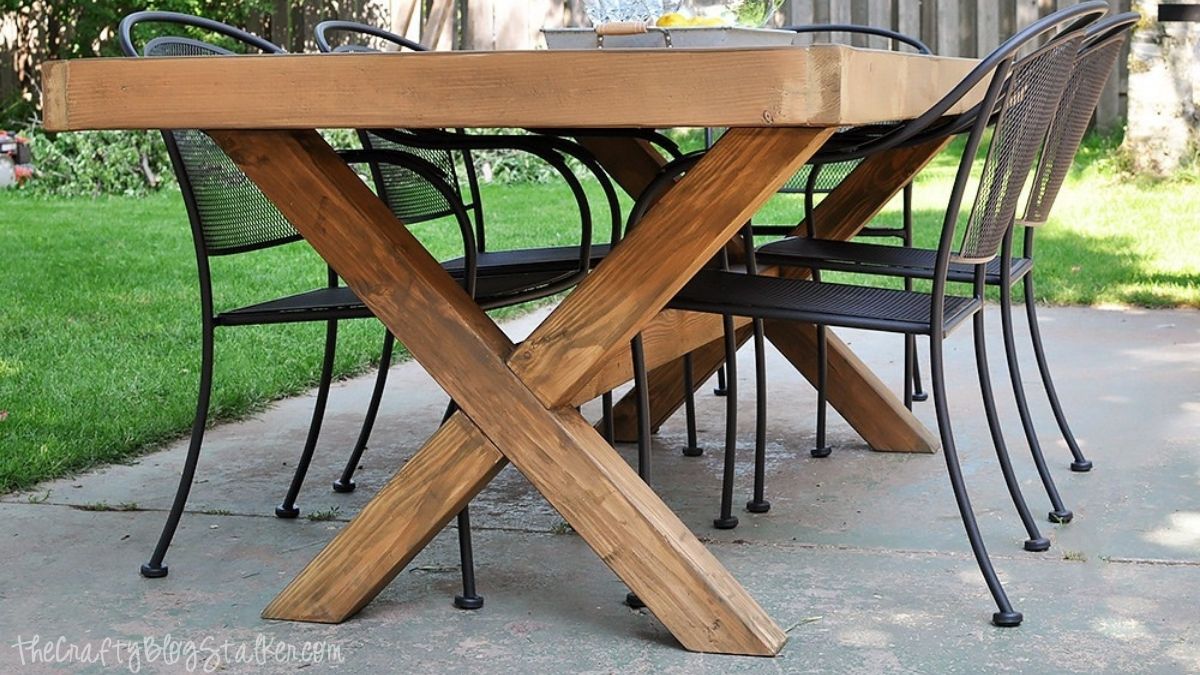 DIY outdoor dining table with a thick wooden tabletop and X-frame base, surrounded by black metal patio chairs on a backyard patio.
