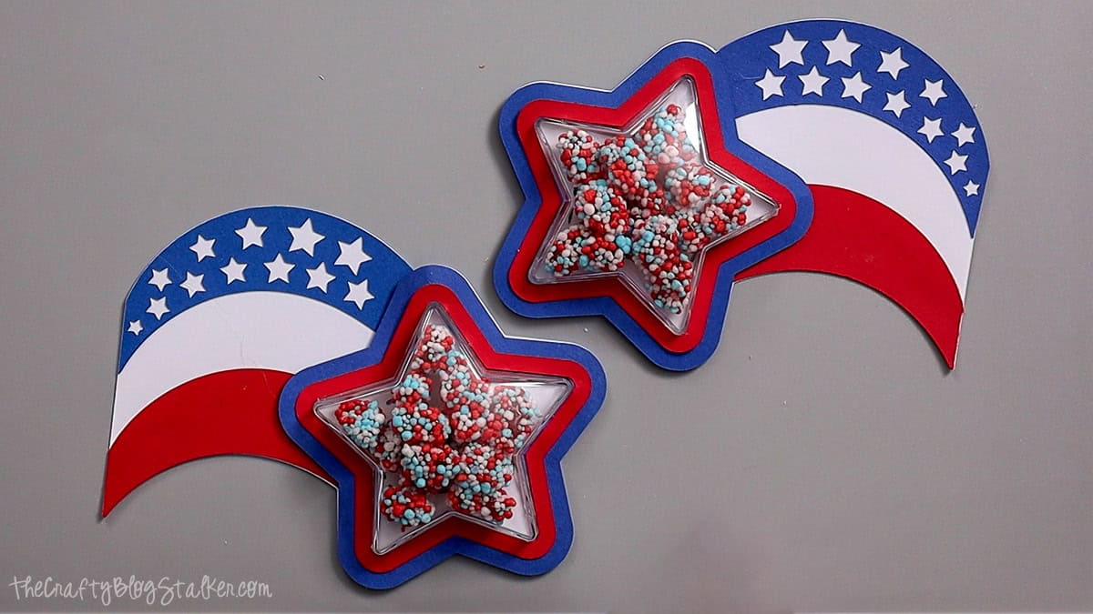 Two patriotic shaker party favors with star-shaped candy containers filled with red, white, and blue sprinkles on layered cardstock.