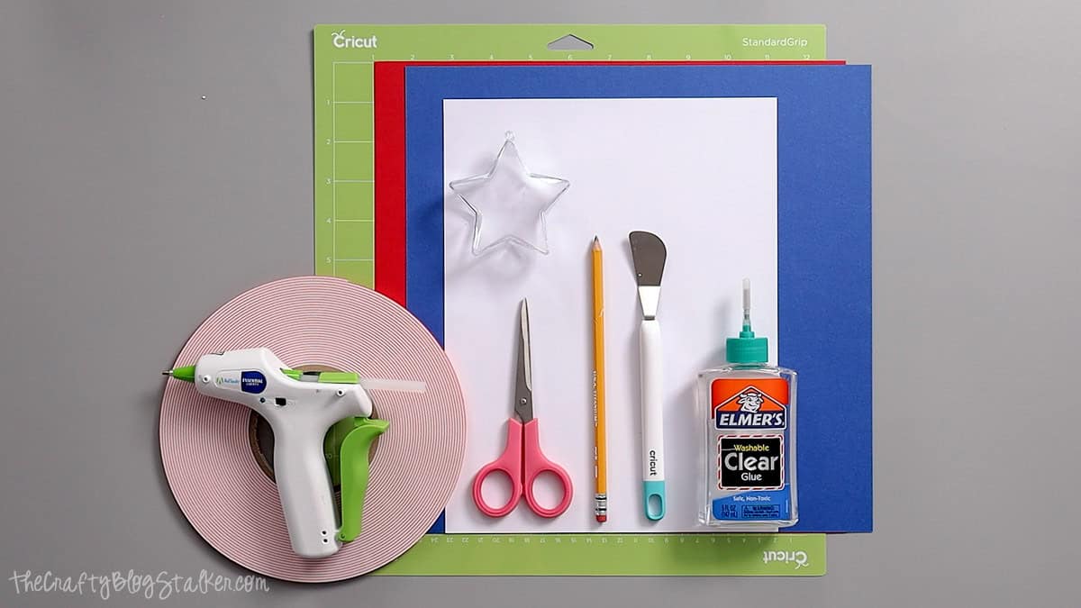 Supplies for patriotic party favors including cardstock, glue, scissors, ribbon, a glue gun, and star-shaped plastic ornaments on a Cricut mat.