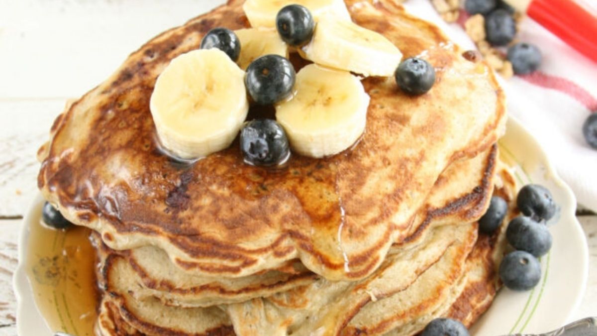 Stack of banana blueberry pancakes topped with sliced bananas, fresh blueberries, and maple syrup on a breakfast plate.