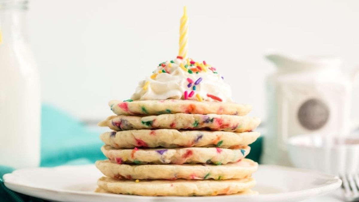Stack of funfetti pancakes filled with colorful sprinkles, topped with whipped cream, rainbow sprinkles, and a birthday candle.